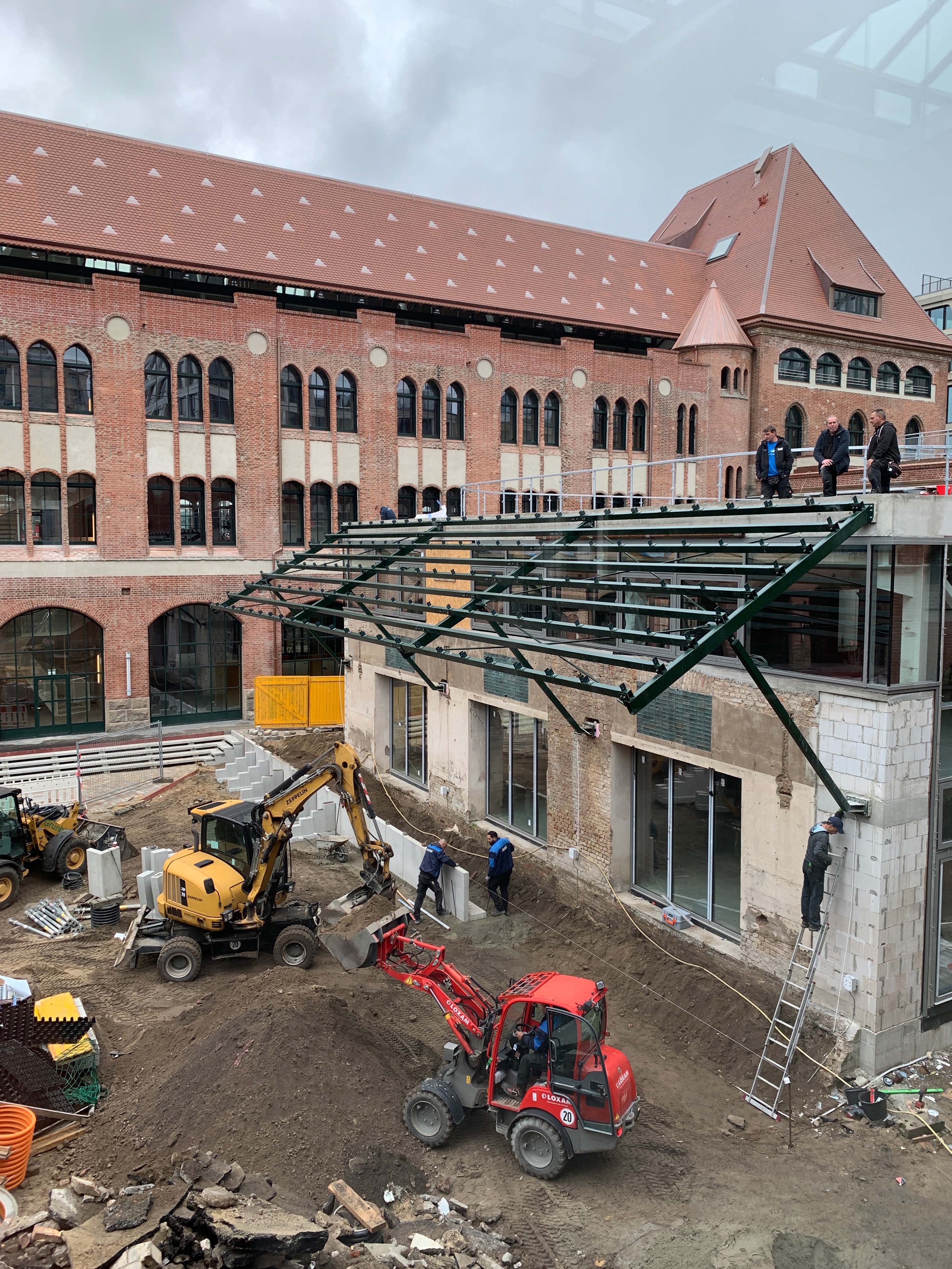 Baustelle vor Backsteingebäude mit Bauarbeitern und Baggern, Blick auf die entstehende Struktur der Packkammer.