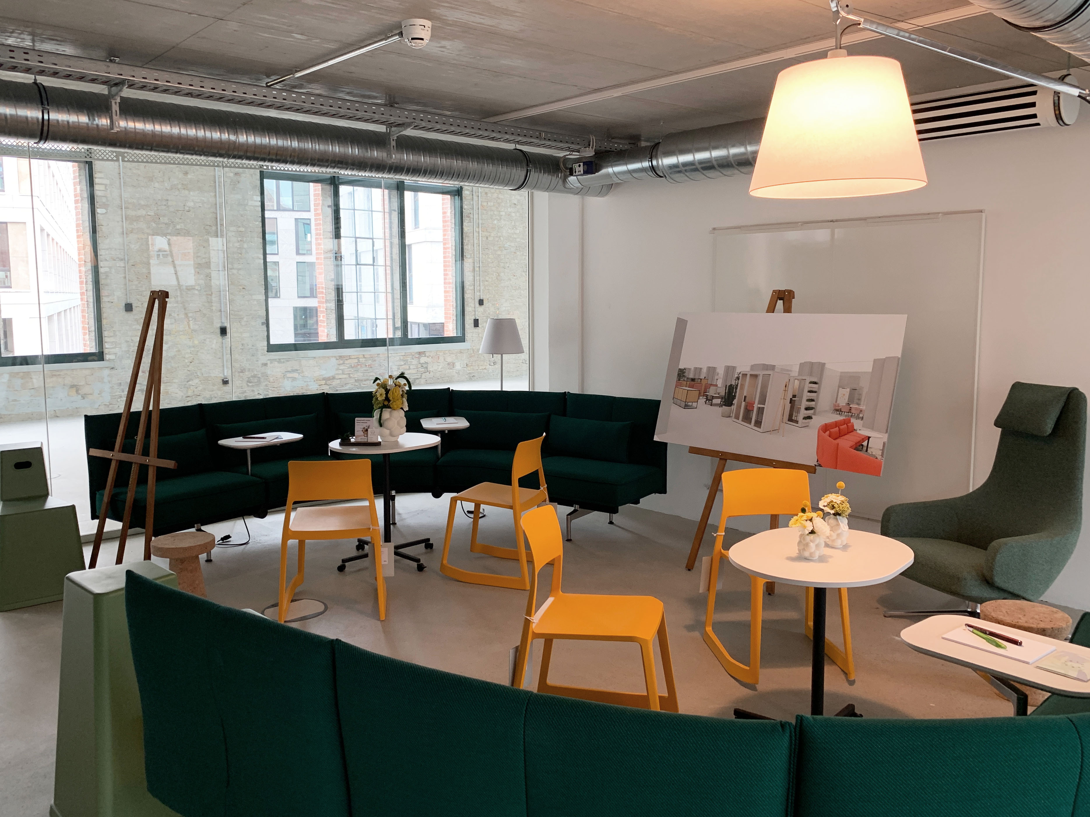 Gemütliche Lounge mit grünen Sofas und gelben Stühlen, eingerichtetem Besprechungsbereich mit Modelltafeln und Stehleuchten.