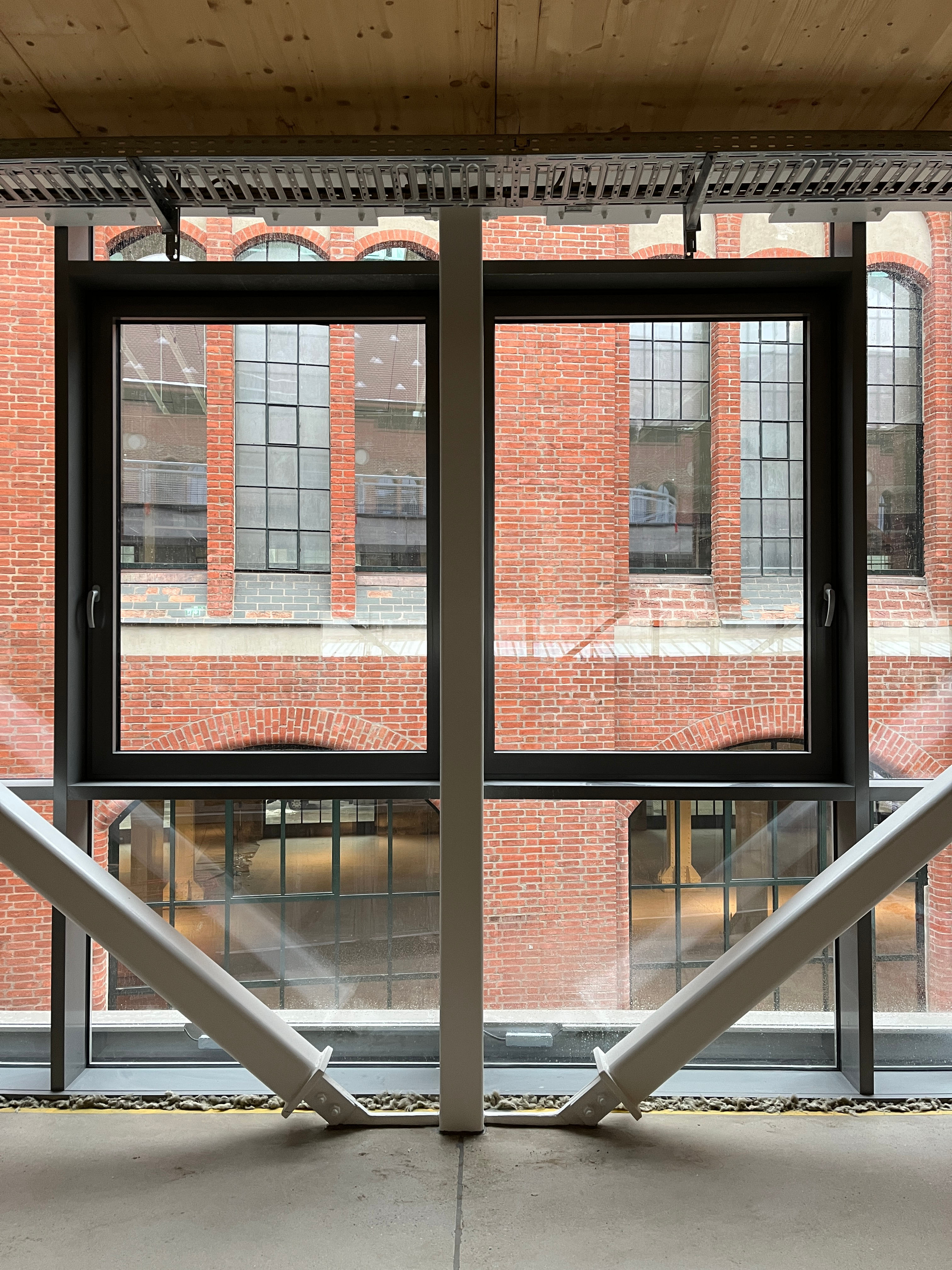 Der Baufortschritt im Postbahnhof Berlin zeigt den Beutel im ersten Obergeschoss mit Blick durch großflächige Fenster mit Metallrahmen auf eine gegenüberliegende Backsteinfassade.