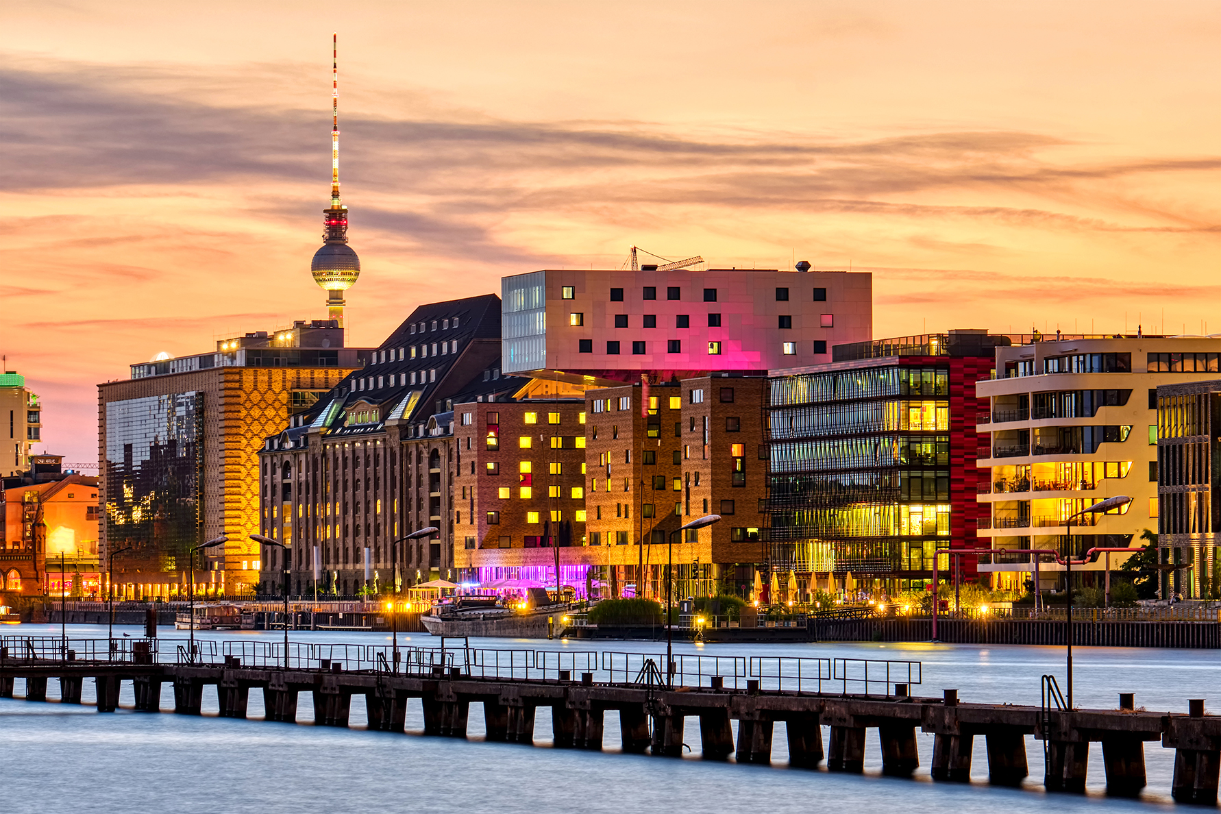 Spreeufer in Berlin mit beleuchteten Gebäuden und dem Fernsehturm im Hintergrund.